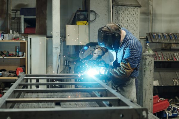 Side view of young male welder in protective face mask carrying out repair or construction work while bending over metallic workbench
